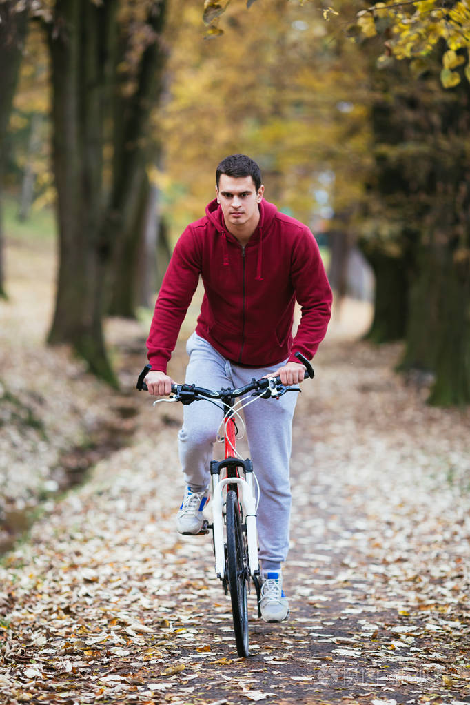 秋季户外活动。年轻严肃英俊的家伙用耳机骑他的自行车在城市公园和听音乐