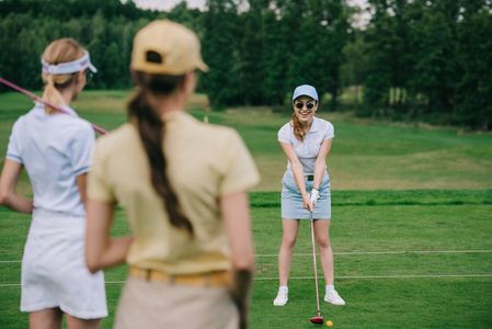 高尔夫器材帽中女性的选择性聚焦看着朋友在高尔夫球场打高尔夫球