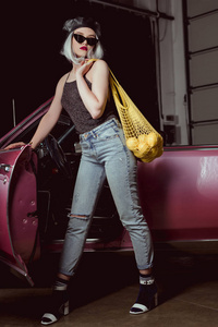 戴贝雷帽和太阳镜的时尚金发女郎的全长视图手持带柠檬的绳子袋, 同时站在老式汽车旁边