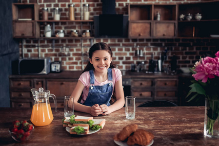 微笑的女孩的肖像看着相机, 而坐在厨房的餐桌上, 在厨房的早餐