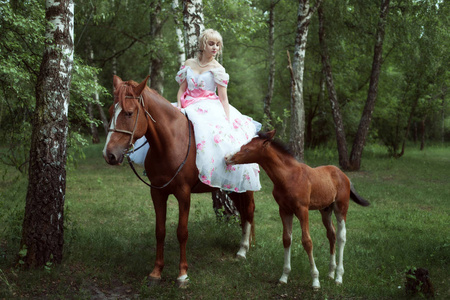 穿着复古连衣裙的漂亮女人坐在一匹马上。他们旁边一只马驹
