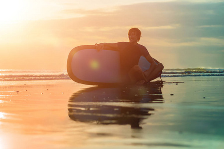 年轻冲浪男子的剪影坐在海边冲浪板在日落时间