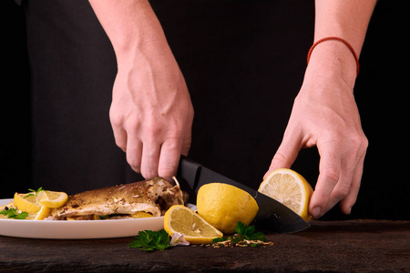 在厨房用木板为烤鱼切柠檬的女人, 手特写。用柠檬香料和香草制成的白盘子煮鱼