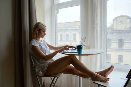 年轻迷人的女孩坐在大窗户旁边的咖啡和工作的笔记本电脑