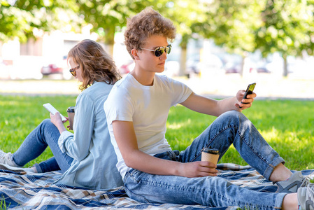 年轻夫妇在爱男孩和女孩。夏天在自然。他们坐在毯子上。放学后他休息。在眼镜的手中, 茶或咖啡。他们看电话。放学后享受放松