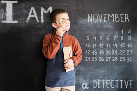 年轻男孩站在与放大镜和笔记本在他的手靠近黑板。年轻的侦探。2019日历的创意设计理念。十一月