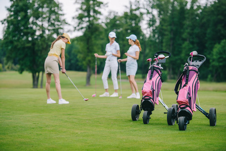 高尔夫球装备的选择焦点, 妇女在帽子与高尔夫球俱乐部和朋友站立在附近在高尔夫球场