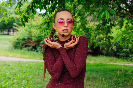 美丽的年轻女子, 长辫子, 粉红色的眼镜和樱桃耳环手捧樱桃