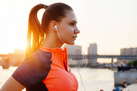 一个年轻的体育健身女孩做运动, 听音乐在耳机的背景下, 通过太阳