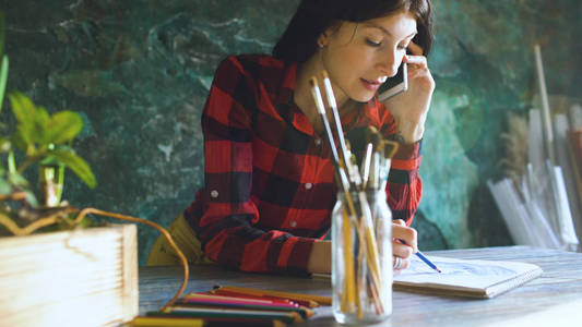 年轻女子画家画 scetch 在纸笔记本上的铅笔和通话电话在室内