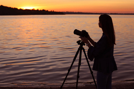 年轻的女摄影师调整专业相机在三脚架在日落