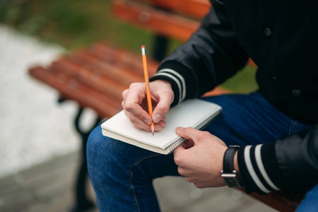 穿着黑色夹克的学生坐在长凳上的公园里, 把他的想法写在笔记本上。英俊的男孩
