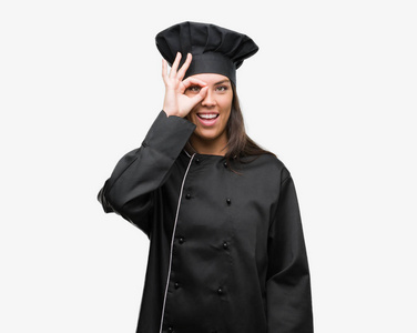 年轻的西班牙裔厨师妇女穿着厨师制服与愉快的面孔微笑做 ok 标志用手在眼睛看通过手指