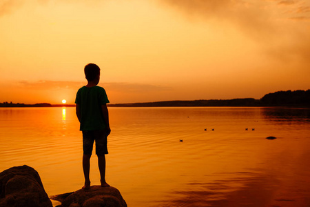 小男孩发射纸船只从河边。在远方的浮船。美丽的夕阳。折纸。河里。湖
