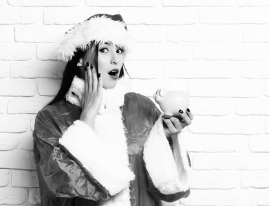 可爱的圣诞老人女孩或惊讶的黑发女人，在新的一年毛衣和圣诞节或圣诞帽子持有的背景墙上的白砖粉色小猪猪银行