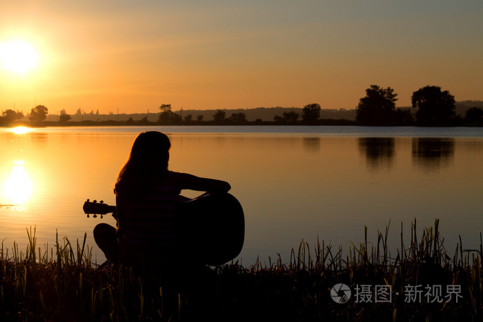 一个女孩弹吉他在河边日落时的剪影