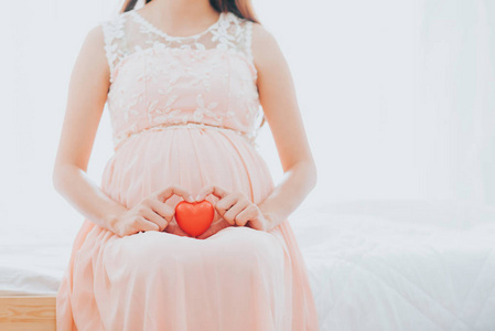 怀孕的妇女抱着红色的心在她怀孕的腹部护理在床房