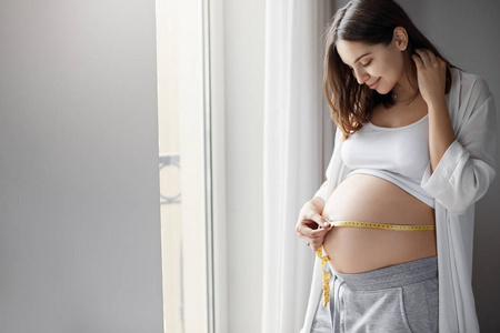 年轻漂亮怀孕女孩测量她的肚子，用胶带来跟踪她的胎儿的发展。健康怀孕概念