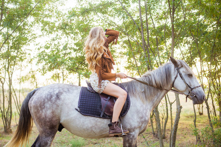 美丽的年轻女孩, 长着金色的头发在一个麂皮绒夹克与条纹微笑, 骑着马在秋季日落