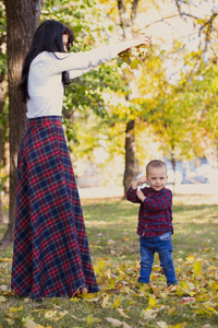 秋天, 美丽的年轻母亲在公园里和儿子玩耍。他们在玩干落叶的时候很开心。