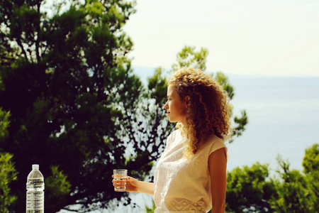 漂亮的女人或卷曲的金发女孩站与玻璃的饮用纯净水和阳台上的塑料瓶在自然背景上的夏天