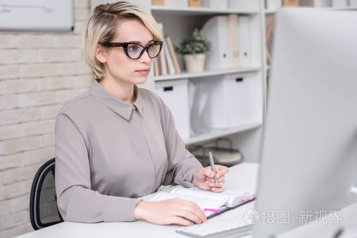 金发女实业家的肖像使用计算机为工作坐在办公桌在现代办公室和写笔记
