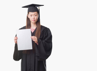 年轻的中国妇女穿毕业制服持有纸学位与自信的表达对聪明的脸思维严重