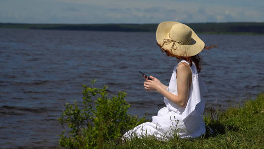 漂亮的红发女孩穿着白色夏季使用一部手机，坐在岸边和河流