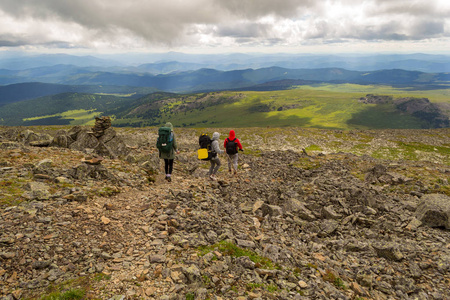 三名游客两个女孩和一个背包的男孩沿着山上的岩石走向绿色的田野, 冒险下的蓝天和云在阿尔泰山命名 sarlyk