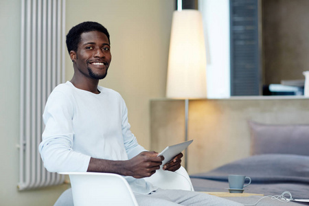 快乐的非洲裔美国自由职业者的肖像看着相机与广泛的微笑, 而在启动项目与数字平板电脑的帮助下, 背景舒适的卧室内部