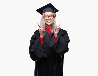 年轻的金发女子穿毕业制服在孤立的背景微笑交叉手指与希望和眼睛关闭。运气和迷信概念