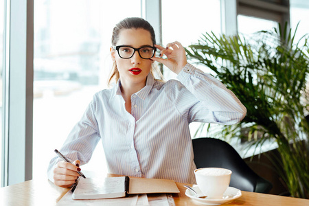 商务女士的肖像在办公室的衬衫和严格的眼镜。这个女人坐在咖啡馆的一张桌子上, 在笔记本上做着