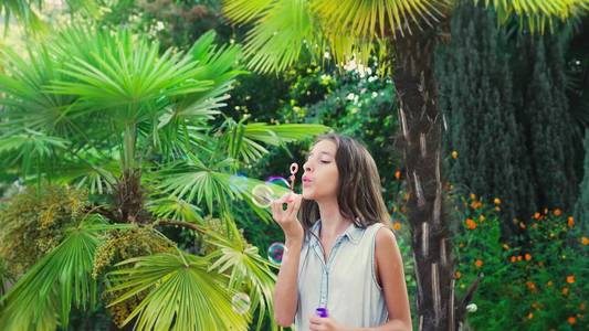 女孩青少年褐发女郎吹肥皂气泡对热带公园背景