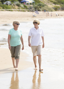 两个可爱的高级退休妇女在他们的60s 有乐趣一起享用愉快的漫步在海滩微笑嬉戏