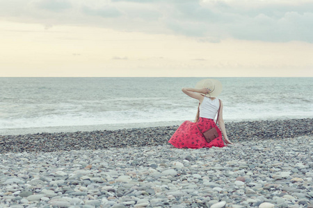 在红色的裙子和帽子的女孩坐在海边，日落的时候。查看从背面