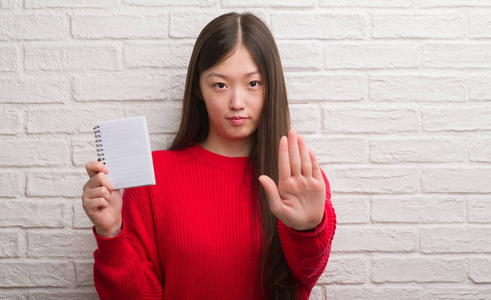 年轻的中国妇女在砖墙上显示空白笔记本用张开的手做停止标志与严肃和自信的表达, 防御手势
