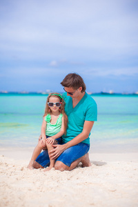 年轻的父亲和他的小女儿坐在热带的白色沙滩上