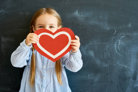 可爱的小女孩的肖像隐藏在红纸心脏背后, 看着相机摆在学校黑板上