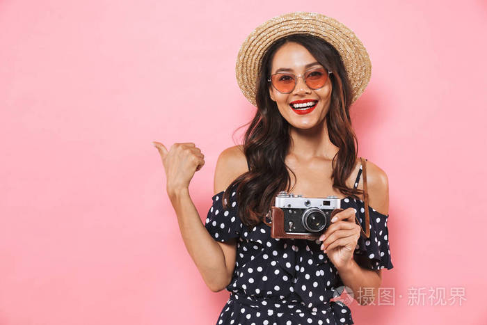 微笑的黑发妇女在草帽和太阳眼镜指向, 而摆在与复古相机和看着在粉红色背景的摄像头