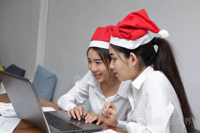 两个开朗年轻的亚洲商界妇女与圣诞老人帽子一起工作的办公场所