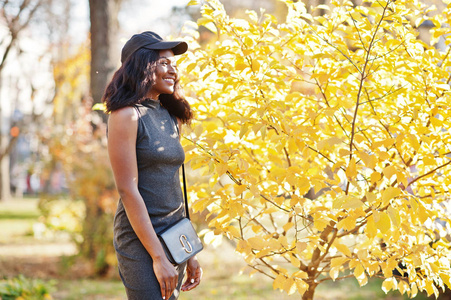时尚的非洲裔美国女孩在灰色的裙衫, 斜挎包和帽子摆在阳光明媚的秋季日对黄叶。非洲模范妇女
