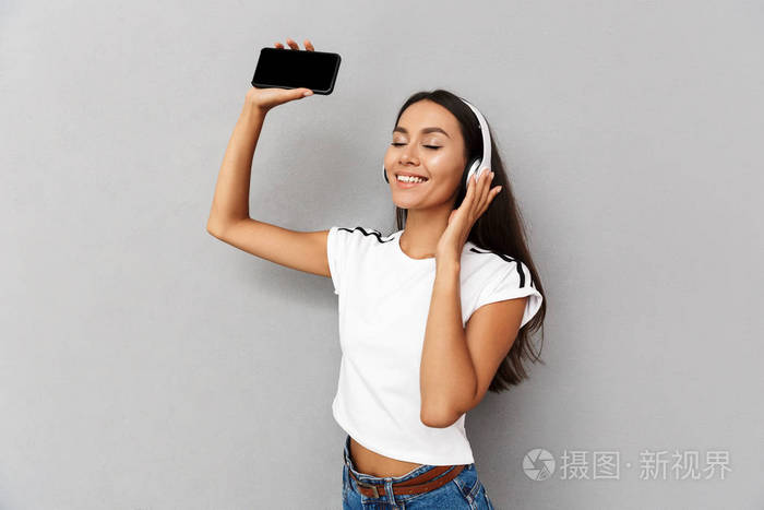 年轻快乐情绪女性的形象用手机听音乐, 耳机被隔离在灰色背景下