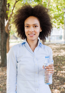 微笑的年轻黑人妇女的画像举行自然瓶水, 非洲头发样式, 穿蓝色衬衣, 看照相机