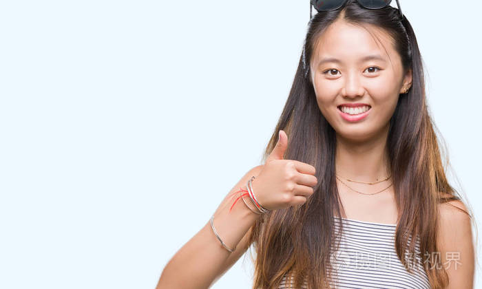 年轻的亚洲妇女戴着太阳镜在孤立的背景下做高兴的拇指手势与手。批准表达式看相机与显示成功