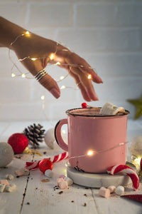 妇女手与搪瓷杯热可可与迷你棉花糖和糖果手杖与美丽的圣诞灯的散景。圣诞理念