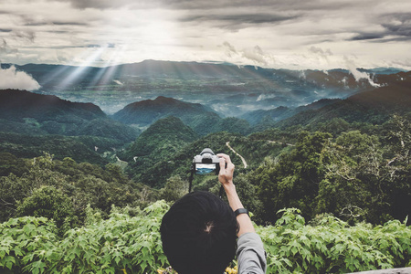享受旅行用的山峰。背包客手里拿一台数码相机在采取与日落山全景照片的山峰光射线