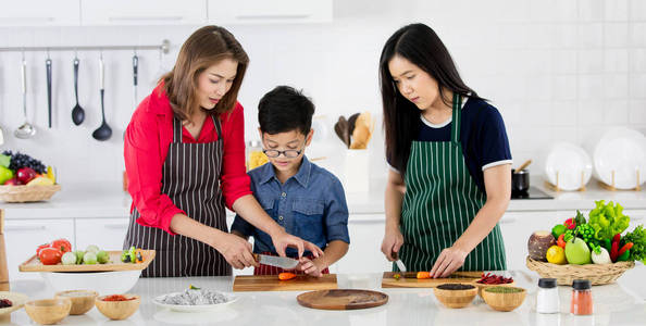 美丽的亚洲妇女在红色衬衫和黑色围裙教她的女儿和儿子如何准备蔬菜烹饪在白色干净的现代厨房