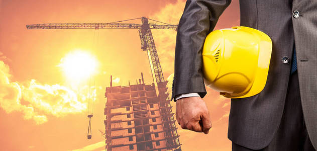 工程师手持黄色头盔在新建筑和建筑起重机背景下, 在黄昏多云的天空下