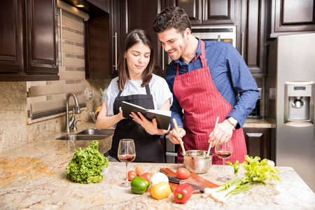西班牙裔夫妇的肖像在围裙读一本烹饪书, 而在厨房准备食物