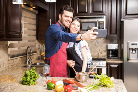 有吸引力的年轻西班牙裔夫妇采取自拍与智能手机, 而在家里做饭的厨房里的食物